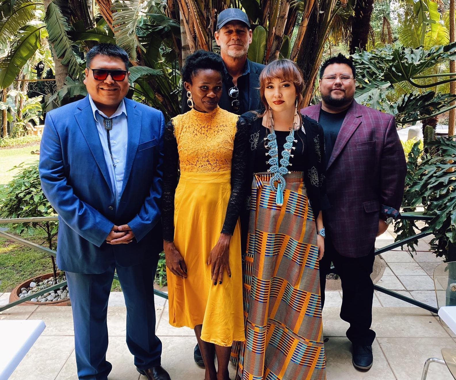 05_DDAT-with-Alex-Rose-and-Neli.jpg A group of five people standing outdoors, dressed in formal and vibrant attire, reminiscent of Ndebele cultural patterns. Lush foliage and a pathway are visible in the background.