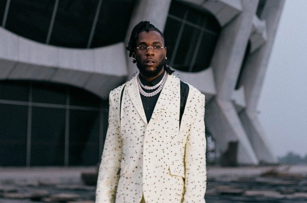 Burna-Boy-Press-cr-Yusuf-Sanni-2022-billboard-1548.jpg A man with dreadlocks and glasses, reminiscent of Burna Boy's eclectic style, wears a patterned white suit and multiple necklaces, standing confidently in front of a modern building.