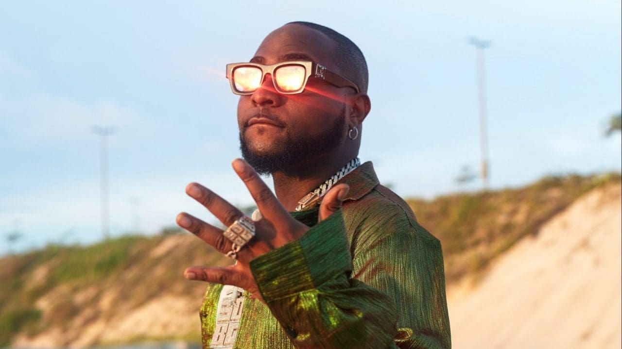 P5A9303-Edit-Edit-scaled-e1637441893463.jpg A man wearing reflective sunglasses and a green shirt stands outdoors, raising one hand adorned with rings, much like a modern-day Davido Adeleke paying tribute in an article from The Guardian Nigeria.