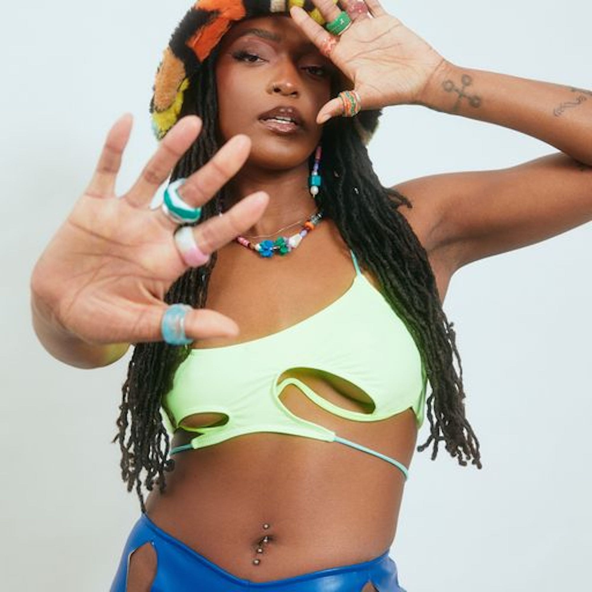 TLbk1lSE.jpeg A woman with long braided hair wearing a lime green cutout top and blue pants poses as if in a music video, holding her hand up to the camera. She wears multiple rings and colorful accessories.