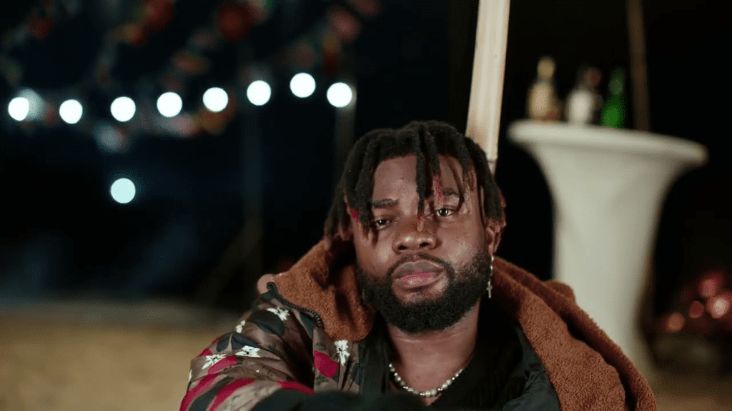 image-30-1024x576-1.png Man with dreadlocks and a beard, wearing a patterned jacket, sits by a white table with bottles in the background. Blurred lights and decorations are visible in the distance, giving off major Senth Music vibes.