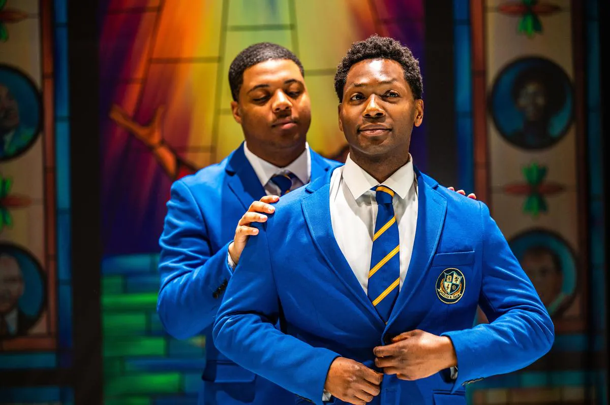 savion_roach_and_andrew_broderick.jpg Two men in blue blazers and yellow-striped ties, one standing behind and adjusting the other's blazer, appear as if preparing for a performance in front of a colorful, stained-glass background. They look like choir boys ready to let their songs soar.