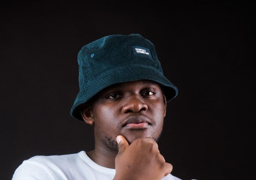 veetchy-drops-stylish-swanky-i.t.m-01.jpg A person wearing a blue bucket hat and white Veetchy shirt poses against a black background, looking directly at the camera with their hand resting on their chin.