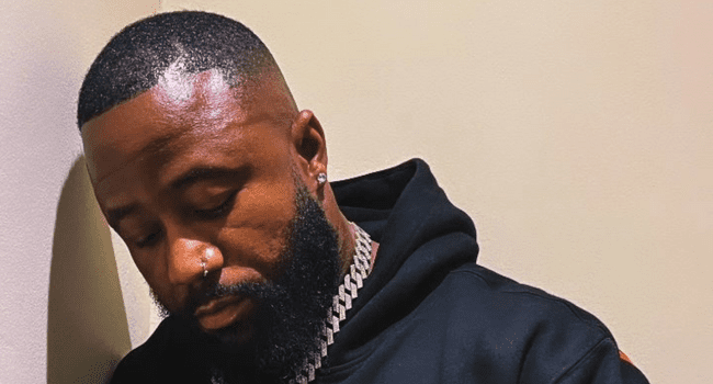 1672446983_Cassper-1.png A man with a beard, wearing a black hoodie and a chain necklace, leans against a beige wall with his eyes closed, embodying the effortless cool of a fellow rich man.