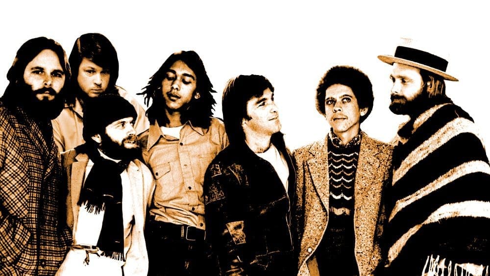 A group of seven men stands together, facing various directions. They are dressed in a mix of casual and formal attire, including hats, scarves, and jackets. The scene evokes an Early '70s vibe reminiscent of the Seaside Boys' "Sail On Sailor" era.