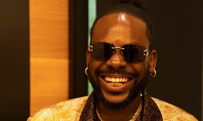 325538073_1258617098332251_4101558424693707621_n-1000x600.jpg A man with sunglasses and earrings smiles broadly, wearing a patterned gold shirt and a necklace, channeling the stylish vibes of Adekunle Gold.