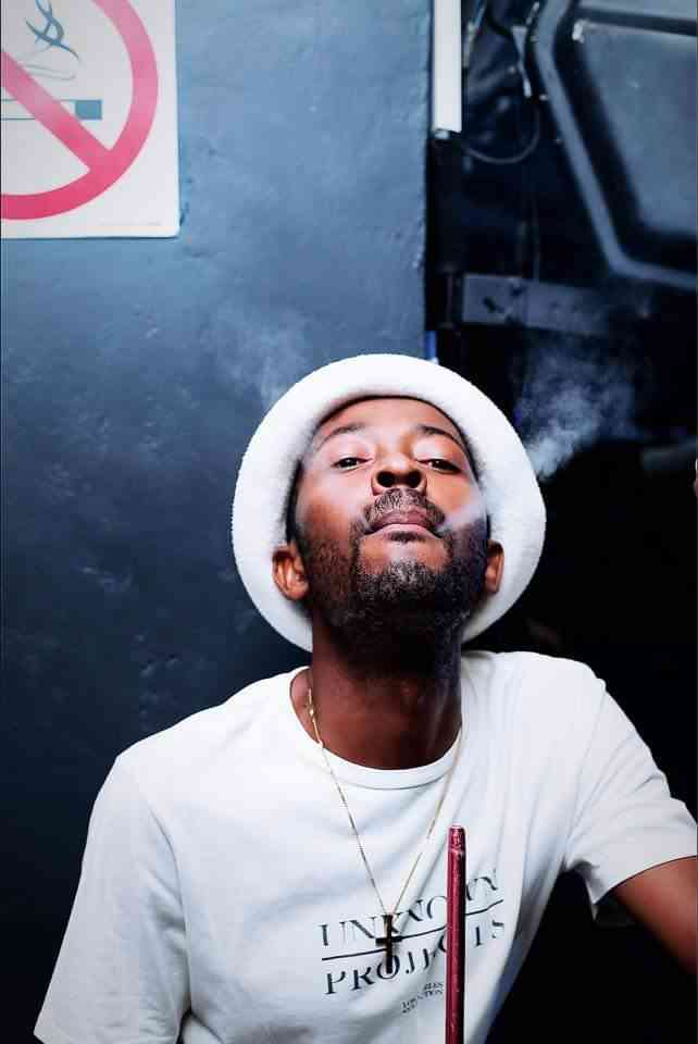 325987969_1894417130918672_1280617090184031605_n.jpg A man wearing a white hat and t-shirt exhales smoke while seated, seemingly oblivious to the "No Smoking" sign visible in the background. His relaxed demeanor resembles Bandros's carefree style.