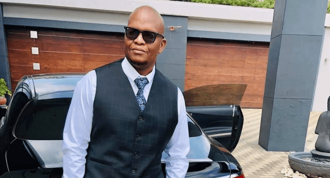 Ambitiouz-entertainment.png A man wearing sunglasses, a white shirt, a patterned tie, and a dark vest stands in front of a black car with a wooden garage door in the background, looking like he could be promoting Sjava's latest new album release from Ambitiouz Entertainment.