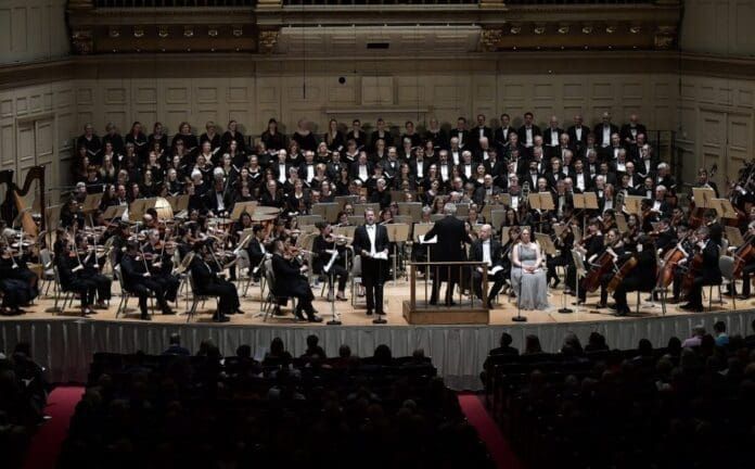 BostonPhilharmonic-ChorusProMusica-1000x620.jpg A symphony orchestra and choir perform on stage with a conductor leading. Musicians play various instruments while a soloist stands near the conductor's podium. A seated audience observes the performance, surrounded by winter decorations that accentuate the classical music experience.