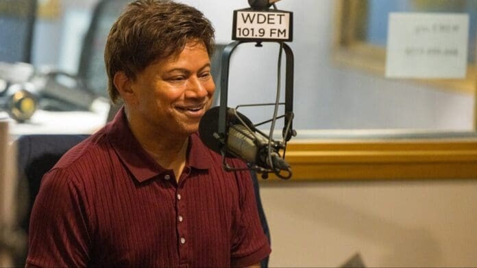 CR5_2889-e1672782734627.jpg A man in a maroon shirt speaks into a microphone at a Detroit radio station with the call sign WDET 101.9 FM visible in the background, discussing black representation on his popular podcast.