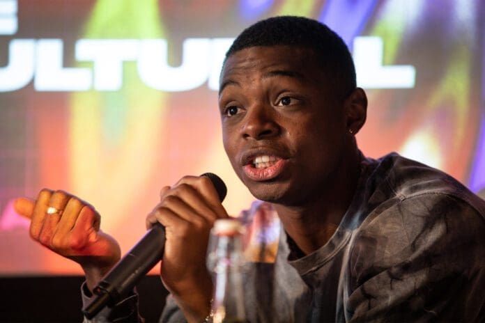 Crooks.jpg Preye Crooks, holding a microphone, speaks at an event with colorful background lights and part of a bottle visible in the foreground.