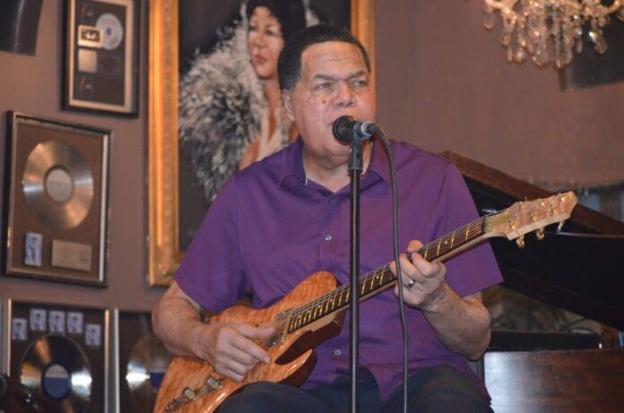 DSC_1957-scaled.jpg A man in a purple shirt plays an electric guitar and sings into a microphone on a stage with framed records and photos on the wall behind him, capturing the essence of blues music.