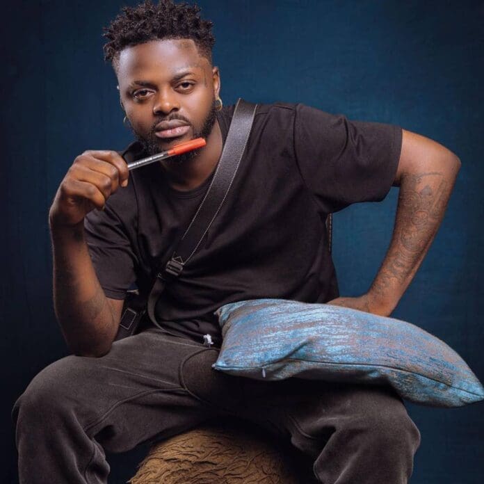 DieselGucci_POW.jpg A man with a beard in a black t-shirt holds a pen near his mouth while sitting. He has a blue cushion on his lap and a dark blue background. The scene exudes the thoughtful vibe often seen in *Passion of the Weiss* interviews.