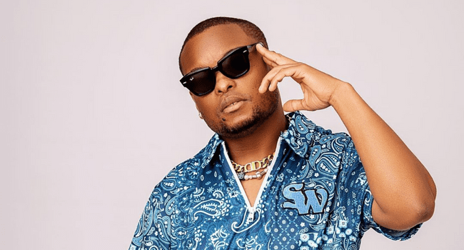 K.O-3.png A person wearing sunglasses, a patterned blue shirt, and a gold chain exudes dominance as they make a gesture with their hand against a light background.