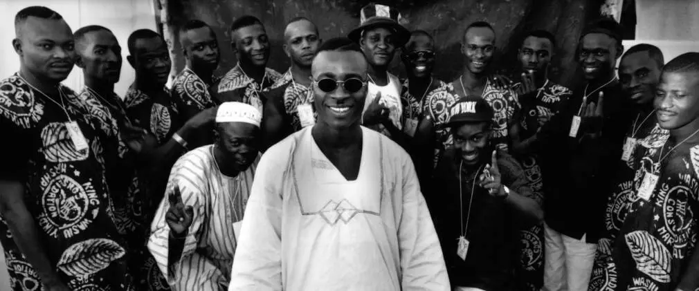 King-Ayinde-cr-David-White-1-e1557841309171-2160x900-1-1010x421.jpeg A group of men, most wearing matching patterned outfits, pose together. One man stands at the front, smiling and wearing sunglasses, embodying the vibrant spirit of Fuji music.
