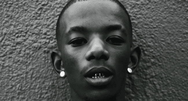 Maglera.png A person with short hair, wearing earrings and sporting a metal dental grill, poses against a textured wall, embodying the edgy vibe of Maglera Doe Boy from the Diaspora Album.