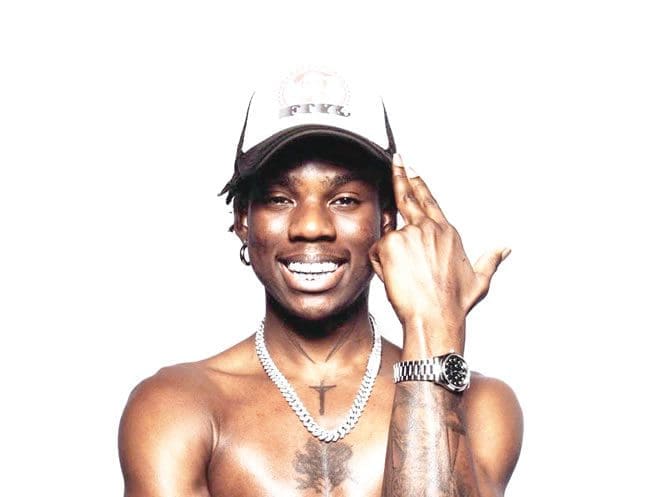 Rema.jpg A man with a bare torso smiles broadly, wearing a baseball cap, chain necklace, and a wristwatch, while gesturing with one hand like the biggest artiste in Benin.