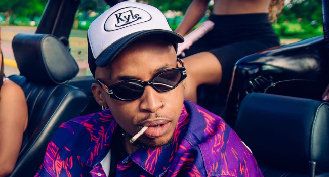 Tshego.png Person in a patterned shirt and sunglasses, wearing a hat labeled "Kyle," sitting in a car with a cigarette in their mouth, jamming to an unreleased rap verse from Tshego.