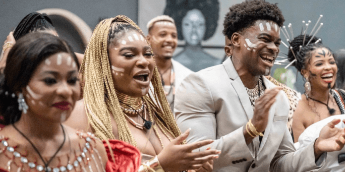 a-still-image-from-inside-the-big-brother-titans-house-of-some-of-the-housemates-smiling-and-laughing.png A group of housemates, dressed in cultural attire and adorned with white face paint, are smiling and clapping at an indoor event, capturing the vibrant spirit that OkayAfrica celebrates.