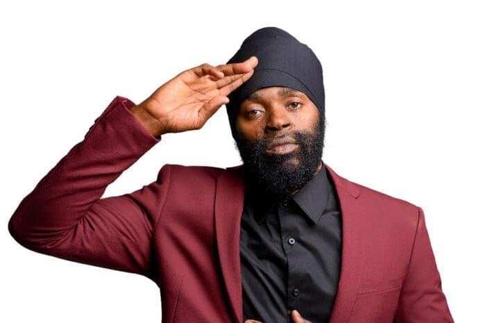 bugle.jpg A man with a beard and black turban, wearing a maroon blazer and black shirt, proudly salutes against a white background. A true DancehallMag moment, reminiscent of Bugle's iconic style.