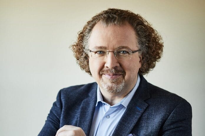 dene_ve_credit_jay_fram_1_.jpg A man with curly hair, glasses, a mustache, and a beard is wearing a blue shirt and a dark blazer, smiling at the camera against a plain background, ready for his interview about conducting the New World Symphony with Stéphane Denève.