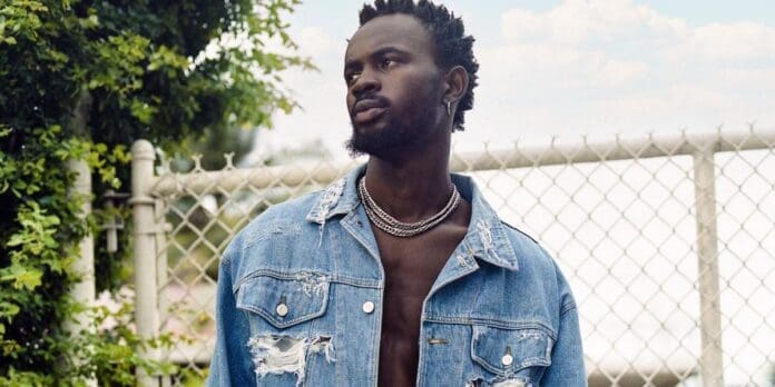 ghana-artist-black-sherif-poses.jpg A man stands outdoors near a chain-link fence, wearing a distressed denim jacket with no shirt underneath, looking to the left. The scene captures the 2023 essence of urban culture that artists like Black Sherif often embody, reminiscent of features on OkayAfrica.