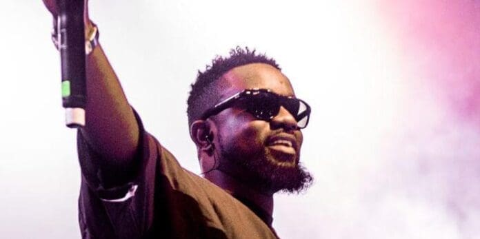 ghanaian-rapper-sarkodie-collabs-with-bob-marley-the-wailers-on-stir-it-up.jpg Man wearing sunglasses holds a microphone and raises his arm, standing in front of a smoky, colorful background, echoing the vibes of Bob Marley.