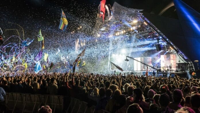 glastonbury_festival_pyramid_stage.jpg A large crowd at an outdoor concert venue at night, with flags and confetti in the air, and a brightly lit stage in the background, reminiscent of the best UK music festivals featured in The Week UK.