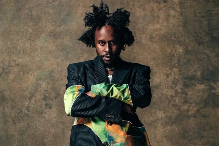 greatishe.jpg Person with crossed arms and a serious expression wears a pinstripe blazer with colorful sleeves, standing against a textured brown background, reminiscent of an album review cover, capturing the essence of Popcaan's "Great Is He.