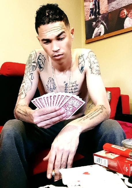 ixa.jpg A shirtless man with tattoos is sitting on a couch holding playing cards. There are card boxes and an open pack of cards on the table in front of him. A picture, possibly portraying a Namibian landscape, is hanging on the wall in the background.