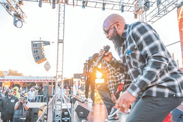 kuJcGirc.jpeg A bearded man in a plaid shirt performs energetically on an outdoor stage, holding a microphone. The stage, set for the latest music, has light and sound equipment, and the audience is visible in the background. Known as Namibian artist Gazza, he captivates everyone with his dynamic presence.