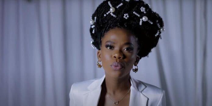 nomfundo-moh-sings-in-her-music-video-for-sibaningi.jpg A person with braided hair decorated with pearls, wearing a white blazer and gold earrings, stands in front of a white curtain during the 2023 OkayAfrica event celebrating African musicians.