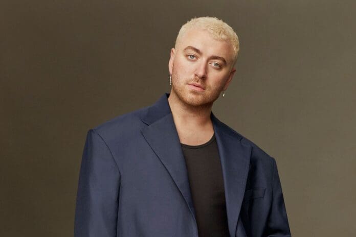 sam_smith_1200_cred_michael_bailey_gates_.jpg A person with short blond hair, reminiscent of Sam Smith's style during his Gloria Tour, is wearing a navy blue blazer and a black shirt, standing against a plain background.