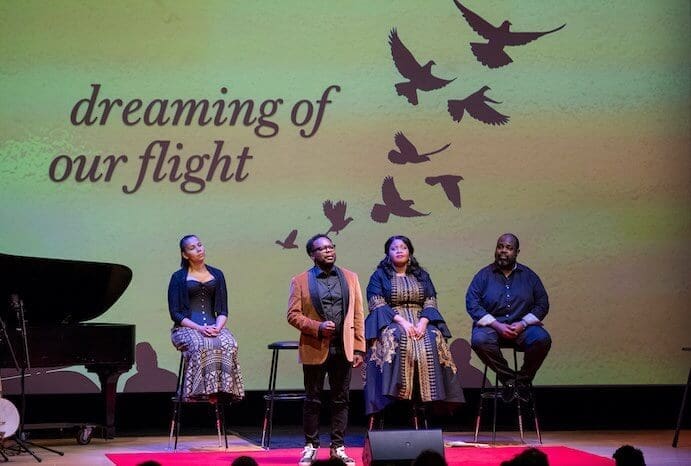 songs-in-flight-photo-by-stephanie-berger-691px.jpg Four individuals are on stage with a piano to the left. Three are seated on bar stools, and one is standing. Background text reads "dreaming of our flight" with bird silhouettes, evoking themes of the African Diaspora in this powerful new song cycle composed by Shawn Okpebholo.