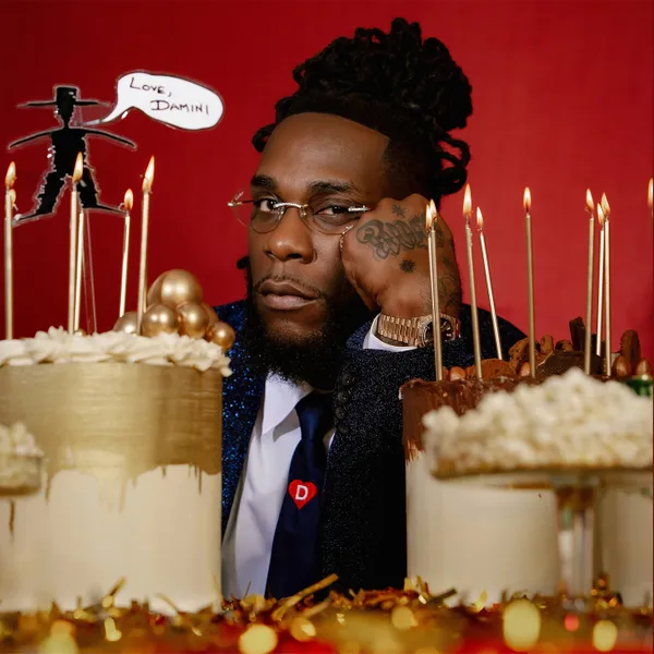 Burna-Boy-Love-Damini.webp.webp A man with glasses and a beard sits behind several cakes with lit candles. He rests his head on his hand with a serious expression, reminiscent of moments in Burna Boy's hit single music video. A speech bubble over him reads, "Love, Damini.