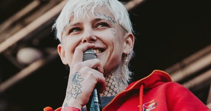 Angie-Oeh_passes_away.jpg Person with short blonde hair and tattoos on their neck and face smiling while holding a microphone, wearing a red jacket. This South African queer rapper, Angie Oeh, captivates the audience with every performance.