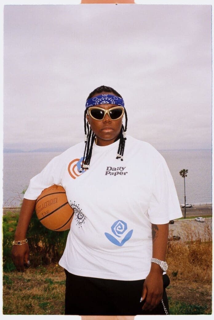 Copy-of-Copy-of-7f3a5758-c9b3-4810-9ee1-55be6e10bbb4.jpg Person wearing a white shirt, sunglasses, and a blue bandana, standing outdoors with a basketball under one arm. There is a beach and ocean in the background, reminiscent of Teni's laid-back vibe as the EQUAL Africa Ambassador for Spotify.