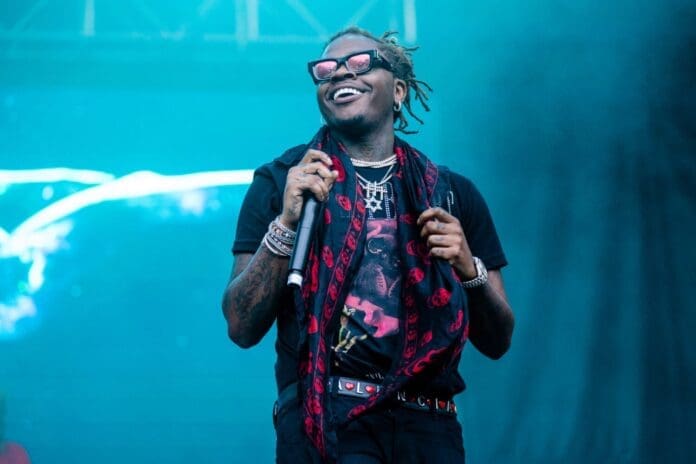 A man wearing sunglasses and a scarf is holding a microphone while promoting Gunna's album "a Gift & a Curse". A man wearing sunglasses and a scarf is holding a microphone while promoting Gunna's album "a Gift & a Curse".