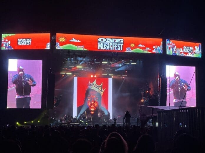 A man on stage at One Music Fest with a large screen in front of him, paying tribute to Hip-Hop stars. A man on stage at One Music Fest with a large screen in front of him, paying tribute to Hip-Hop stars.