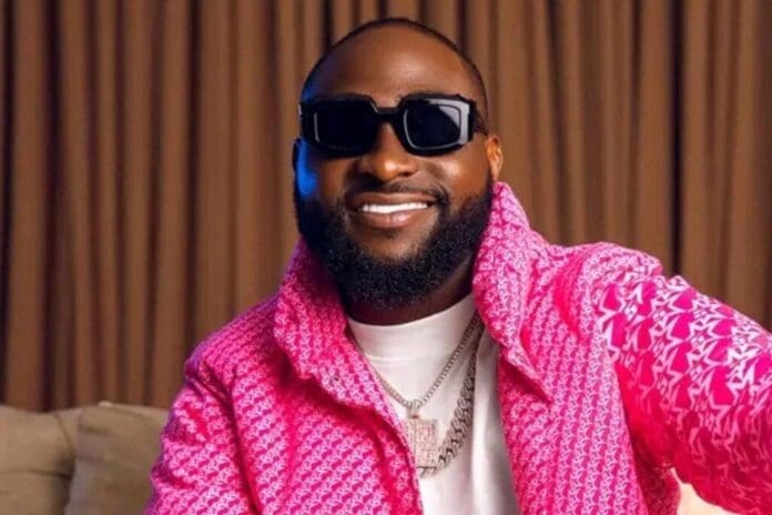 11787572.jpg A man wearing dark sunglasses and a pink patterned jacket smiles at the camera, seated indoors with brown curtains in the background—a scene capturing the vibrant spirit of Afrobeats and global music spotlighted on GhanaWeb.