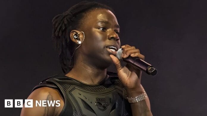 3969d1b0-2d4e-11ef-a044-9d4367d5b599.jpg An individual sings into a microphone on stage, wearing a dark vest, in-ear monitors, and a bracelet. As the BBC News logo glows in the bottom left corner, they captivate the audience with an Afrobeats vibe reminiscent of Rema's "Calm Down.