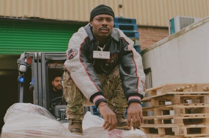 Asake-Press-cr-Juanita-Richards-2024-billboard-1548.jpg A person crouches on bags in a warehouse area, wearing a dark beanie, jacket, and camo pants, reminiscent of an Omah Lay music video. A forklift with a driver is visible in the background, completing the Afrobeats vibe.