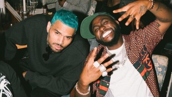 Chris-Brown-Davido-1024x576.jpeg Two men sit together at a crowded table; one has turquoise hair and a black shirt, while the other smiles widely, wearing rings and making hand signs—capturing a vibe reminiscent of Chris Brown and Davido’s dynamic collaborations.