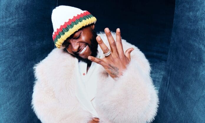 Davido-scaled.jpg A person wears a white fur coat and a colorful knit hat, smiling with eyes closed and showing rings on their fingers against a dark blue background—capturing a vibe reminiscent of Davido’s style.
