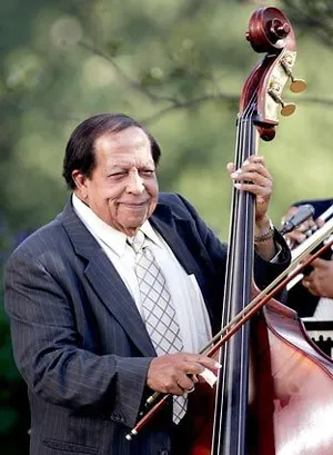 An older man in a suit plays a double bass outdoors with a bow, smiling at the camera.