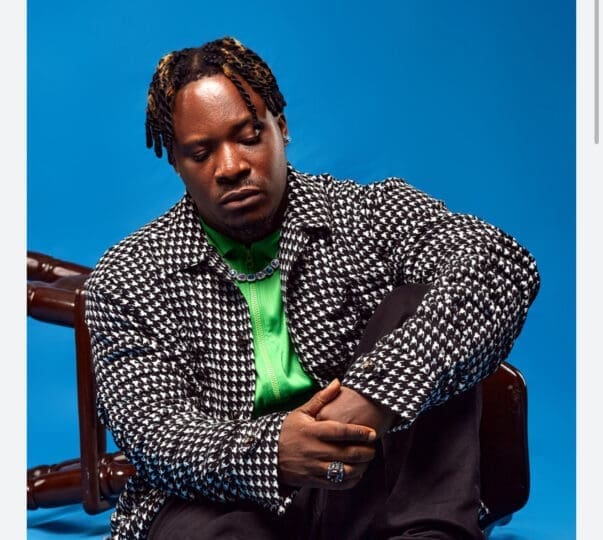Person in houndstooth jacket and green shirt sits against a blue background, looking down, with a contemplative expression.