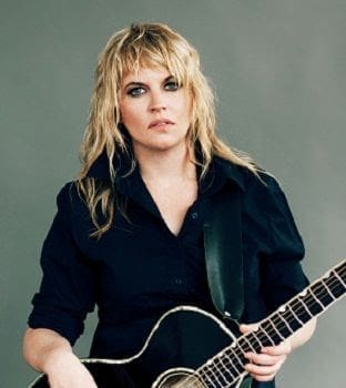 Person with long, wavy blonde hair holding a black acoustic guitar, wearing a dark shirt, gazing at the camera against a plain background.