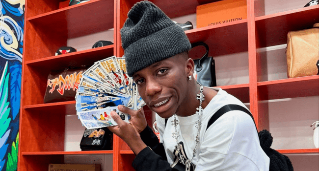 Kindlynxsh-2.png Person wearing a beanie and jewelry, holding a fan of money, stands confidently before shelves adorned with various items. Their expression reflects the proud spirit of Africa's Musical Soldier, embracing dual heritage in a world rich with culture and creativity.