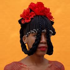 Person with braided hair adorned with red flowers and white beads, standing in front of an orange background.