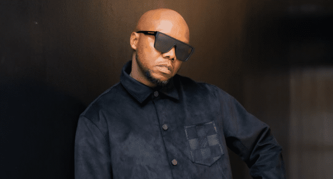 Tbo-Touch.png A person wearing large sunglasses and a dark button-up jacket stands against a dark background, looking at the camera with a neutral expression, exuding the cool confidence of one of the Top 5 SA Rappers.